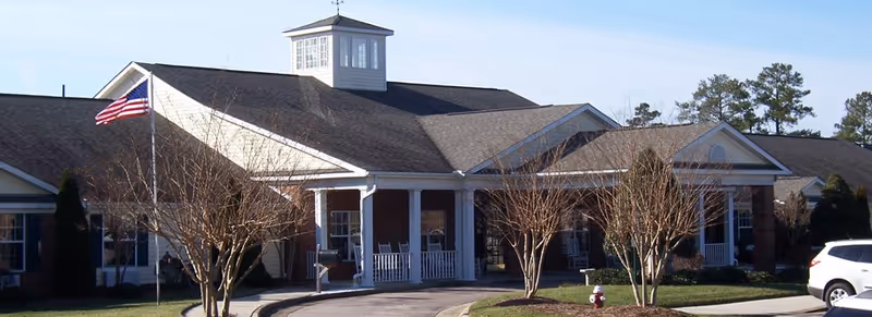 Exterior view of a single-story senior living facility building with a covered entrance, white columns, and a cupola on the roof. There are leafless trees and an American flag on a flagpole in front of the building, along with a driveway and a parked white vehicle.