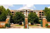 Entrance to Richland Place featuring a driveway flanked by brick pillars with decorative lamps, green signage, and a multi-story residential building in the background surrounded by trees under a partly cloudy sky.