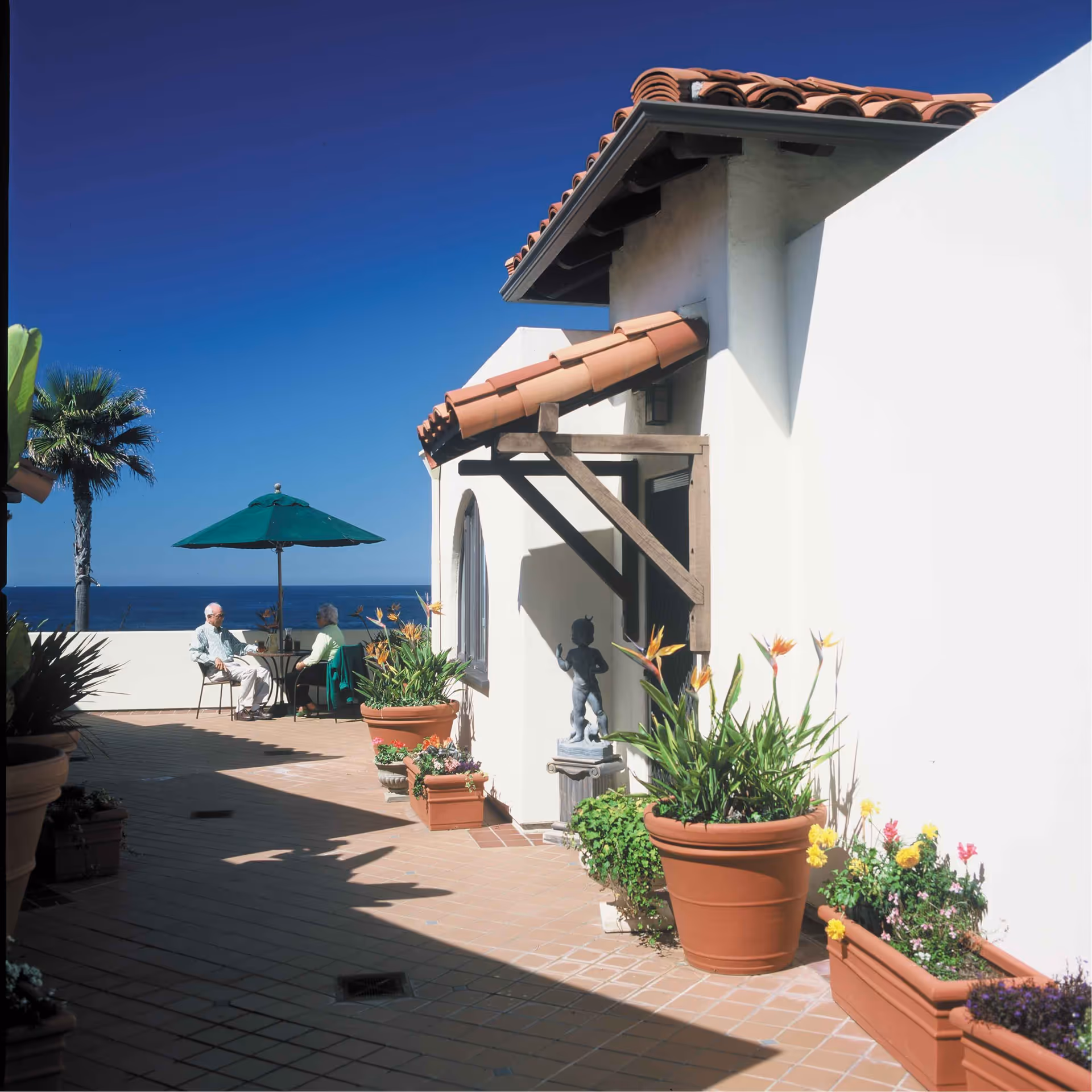 Outdoor patio area at Casa de Mañana Retirement Community with terracotta tiled flooring, potted plants, a small statue, and two elderly people sitting at a table under a green umbrella with a view of the ocean and a palm tree in the background.