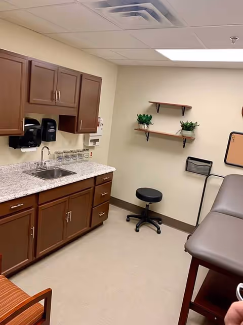 A small medical examination room with a countertop and sink on the left side, brown cabinets above and below the counter, a black rolling stool, two small shelves with potted plants on the wall, a padded examination table on the right, and a chair with orange striped upholstery partially visible in the foreground.