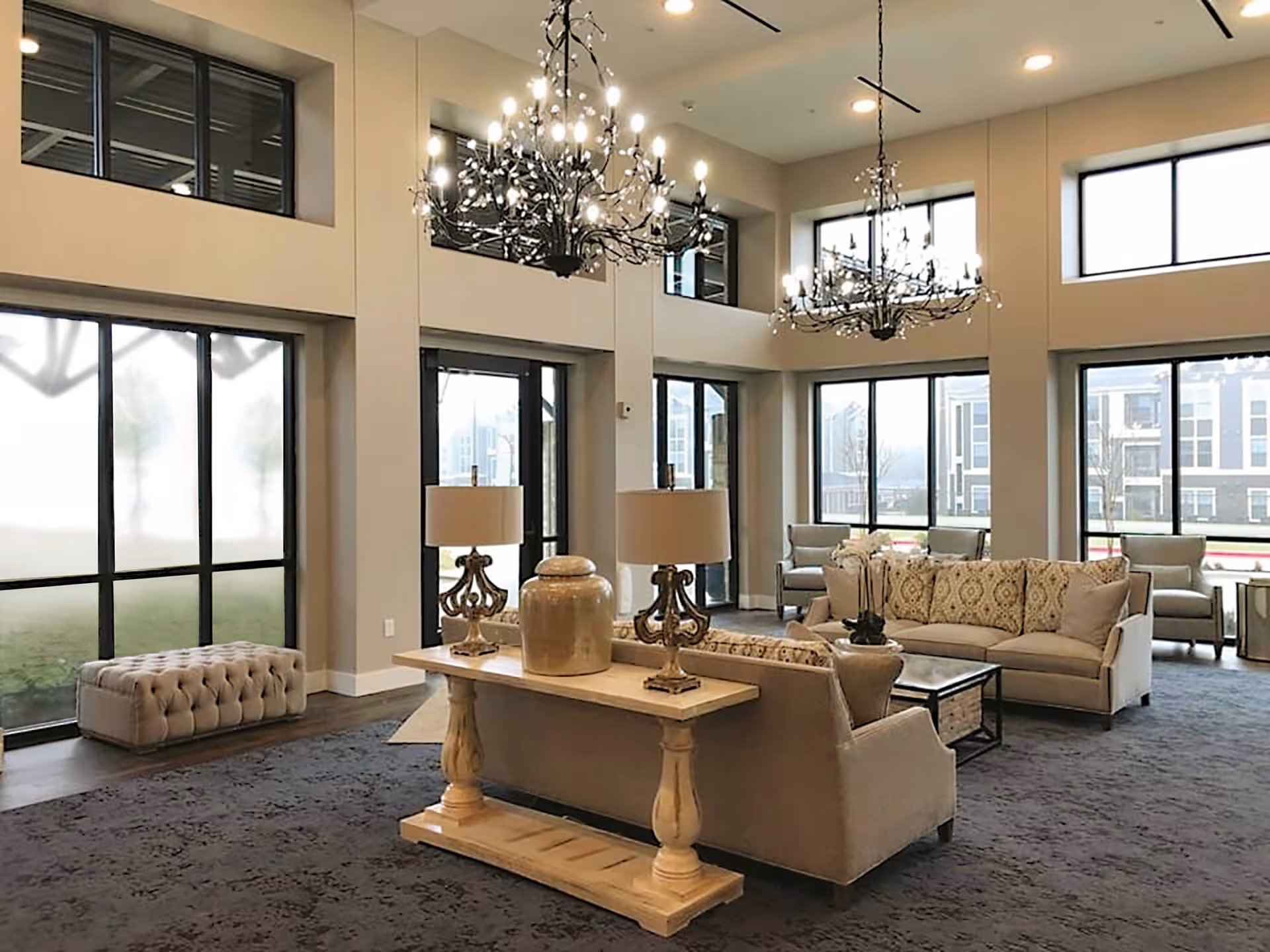 Bright double-height common lounge with sofas, coffee tables, chandeliers, and large windows.