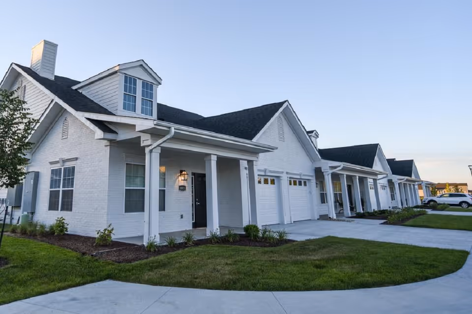 Exterior view of a row of white brick residential buildings with black roofs, each with a garage and a small front porch. The buildings are part of a senior living facility named Traditions at Brookside. The scene is taken during daylight with a clear sky and well-maintained green lawns and landscaping.