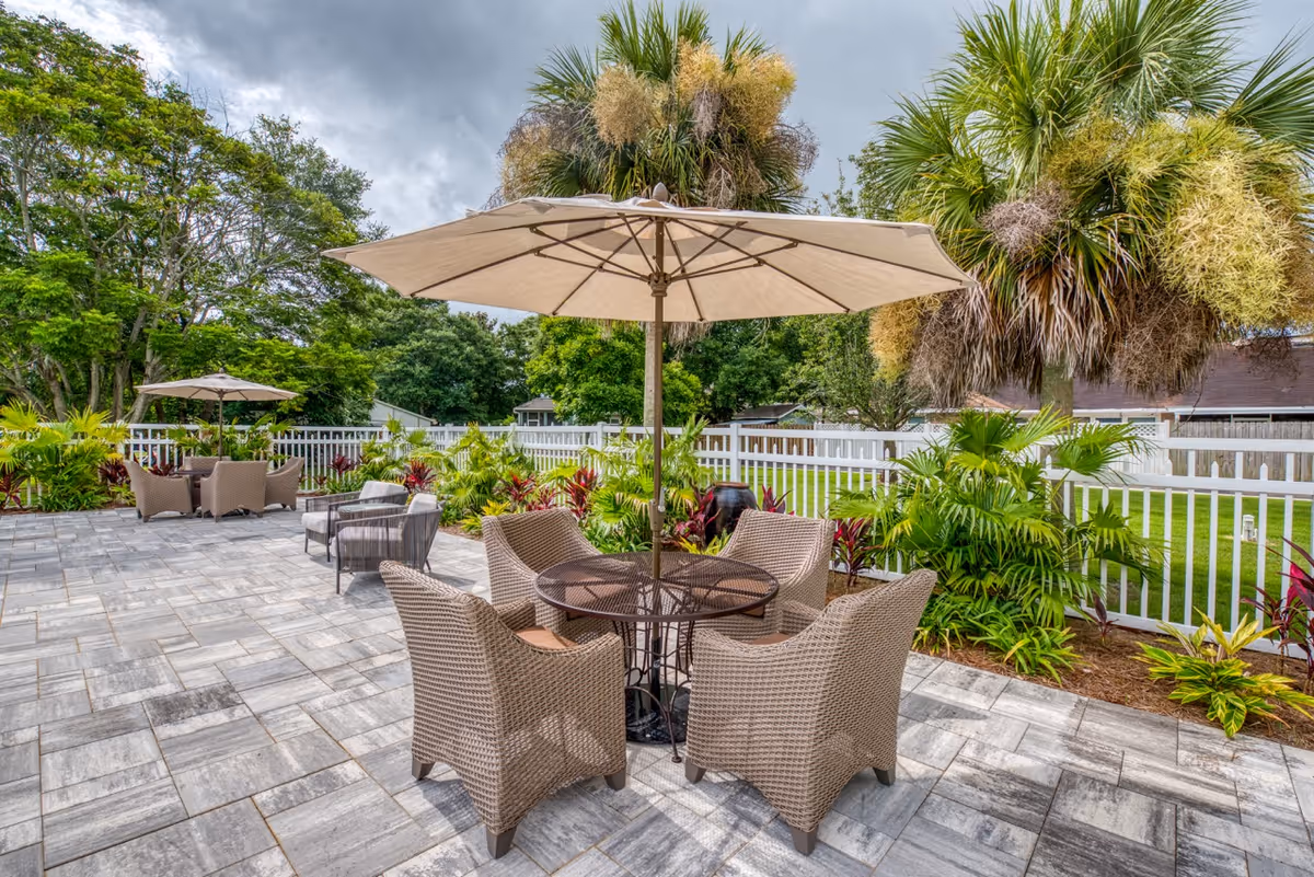 Outdoor patio area with a round table and four wicker chairs under a large beige umbrella. Another seating area with chairs and an umbrella is visible in the background. The patio is surrounded by lush greenery, palm trees, and a white fence.