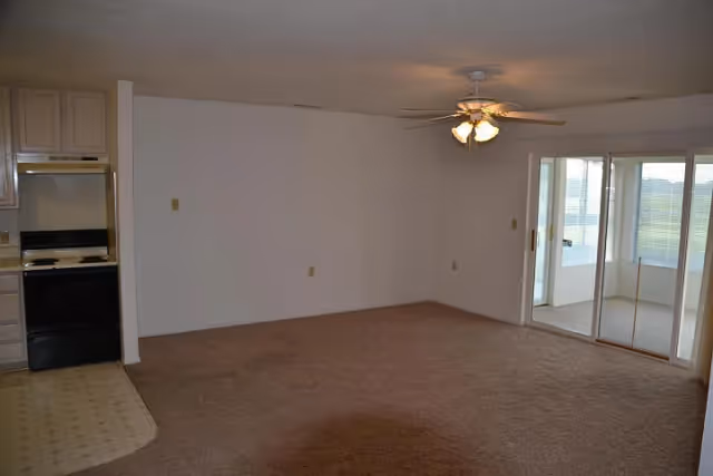 Empty living room area with beige carpet, white walls, and a ceiling fan with lights. To the left is a small kitchen area with a stove and oven. On the right side, there are sliding glass doors leading to a sunroom or enclosed porch with windows and a view outside.
