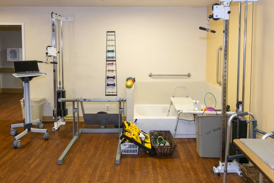 A therapy or rehabilitation room with exercise equipment including a weight machine, a rack of small dumbbells, a laptop on a rolling stand, a bathtub with a shower chair, and baskets containing various therapy tools and equipment. The room has wood flooring and light-colored walls.