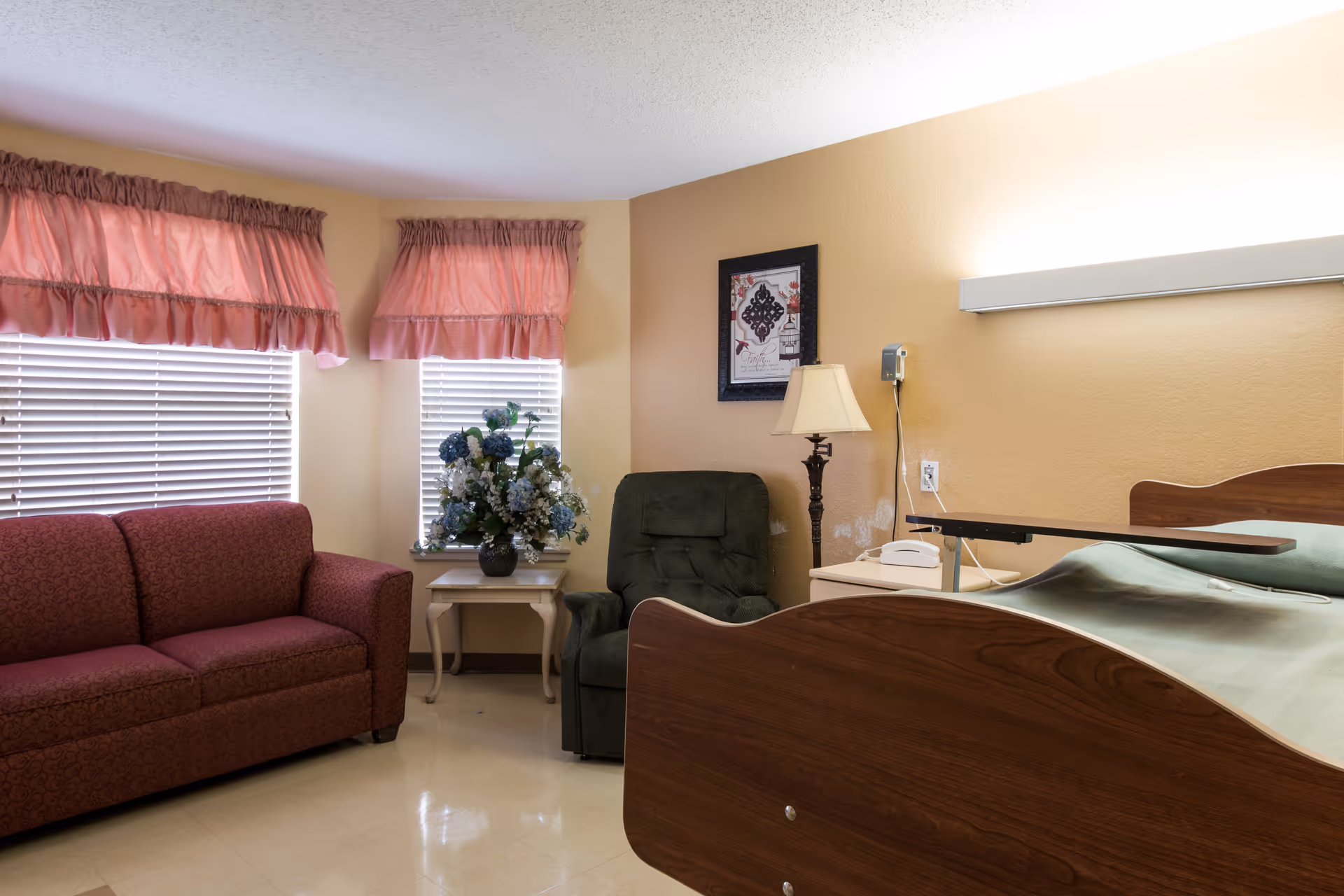 A senior nursing room with a hospital-style bed, red sofa, green armchair, side table with a floral arrangement, lamp and windows with pink valances.