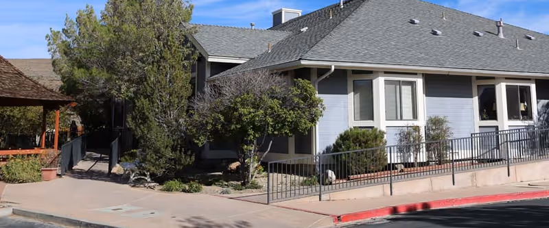 Exterior view of a single-story assisted living facility building with light blue siding, several windows, a gray shingled roof, and a wheelchair-accessible ramp. There are trees and shrubs around the building and a covered walkway on the left side.