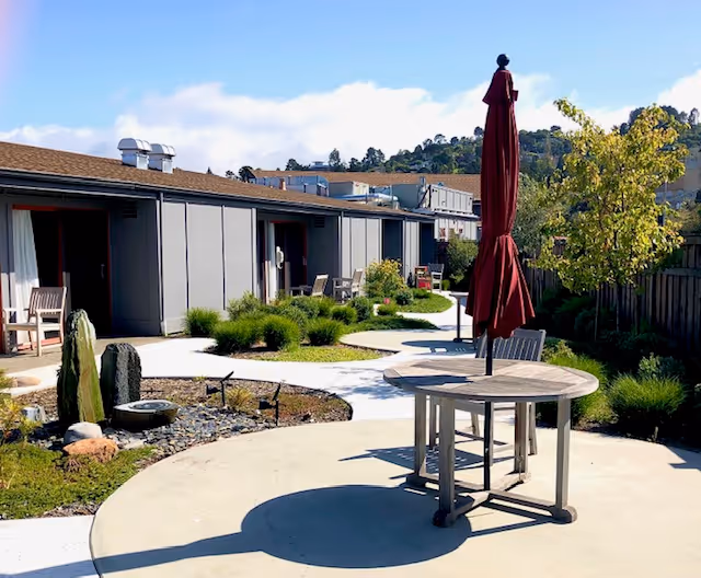 Outdoor patio area at a senior living facility with round tables, chairs, and closed red umbrellas. There are pathways, green shrubs, and a small water feature with rocks. The building has a single-story design with multiple doors and windows, and there are hills and trees in the background under a partly cloudy sky.