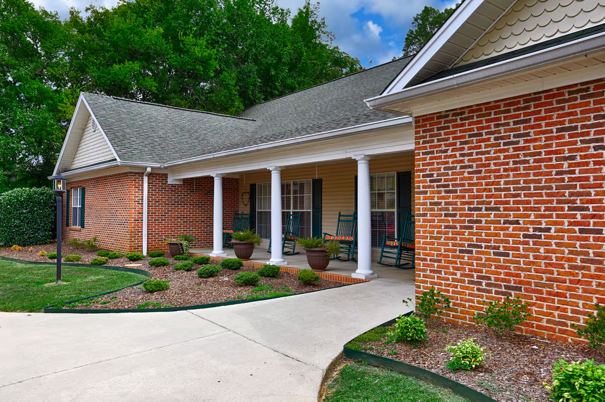 Brick single-story building with a covered porch supported by white columns, green rocking chairs, and a landscaped front walkway.