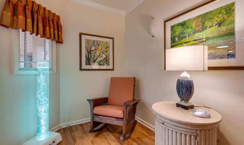 A cozy corner of a room featuring a wooden rocking chair with orange cushions, a round side table with a decorative lamp, a tall illuminated bubble tube light, and two framed paintings on the walls. The room has light-colored walls, a window with blinds and an orange valance, and wooden flooring.