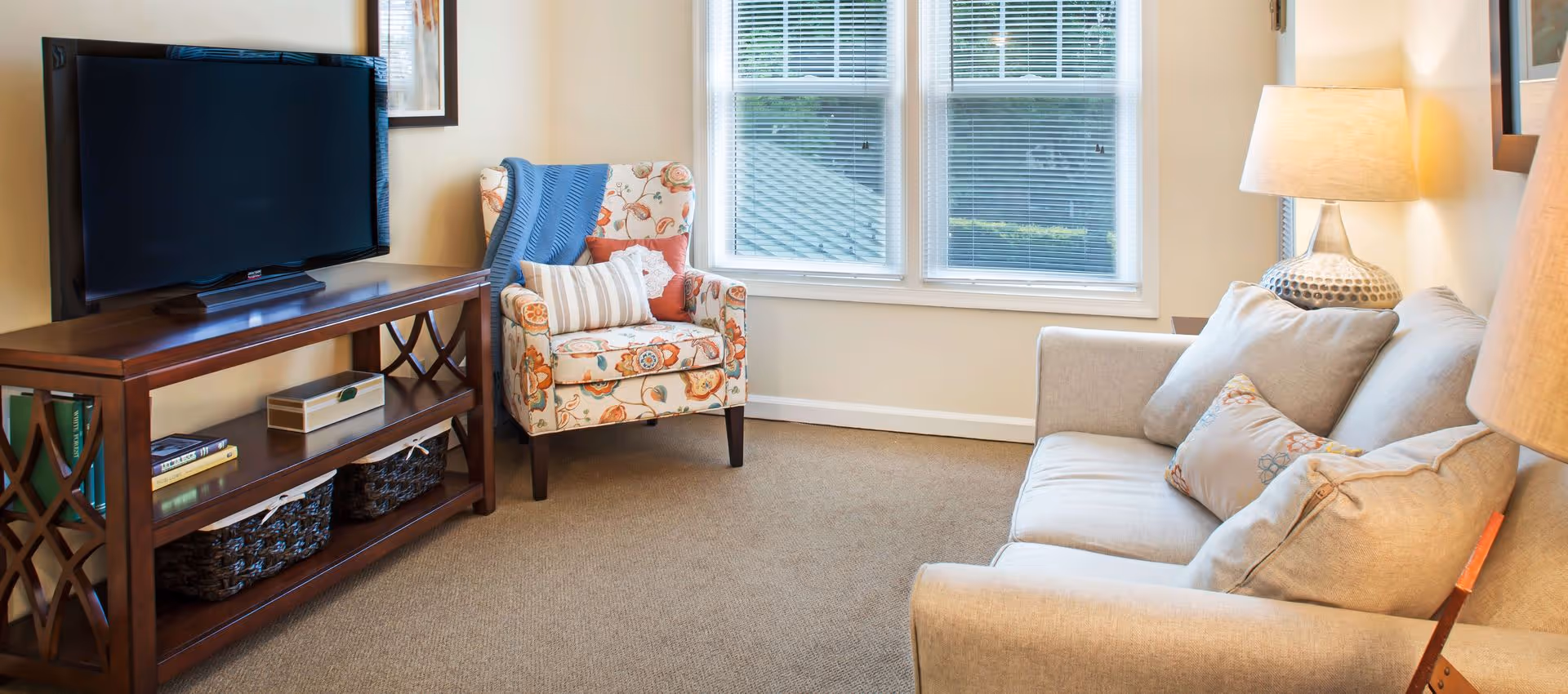 Bright living room with a sofa, patterned armchair, TV on a wooden console, and a lamp by a window.