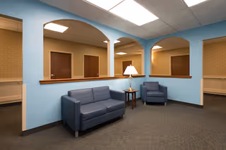 A waiting or lounge area with light blue walls and three large arched openings. The room contains a blue loveseat, a blue armchair, a small wooden side table with a lamp, and a carpeted floor. Several closed wooden doors are visible through the arches.