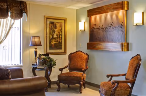 Cozy senior living lounge with two upholstered chairs, lamp, plant, and Thatcher Brook sign.