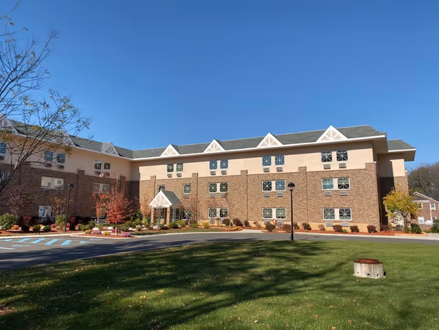 Exterior view of a multi-story senior living facility building with a combination of brick and beige siding under a clear blue sky. The building has multiple windows, a small covered entrance, landscaped bushes and trees with autumn foliage, a paved driveway, and a grassy lawn in the foreground.