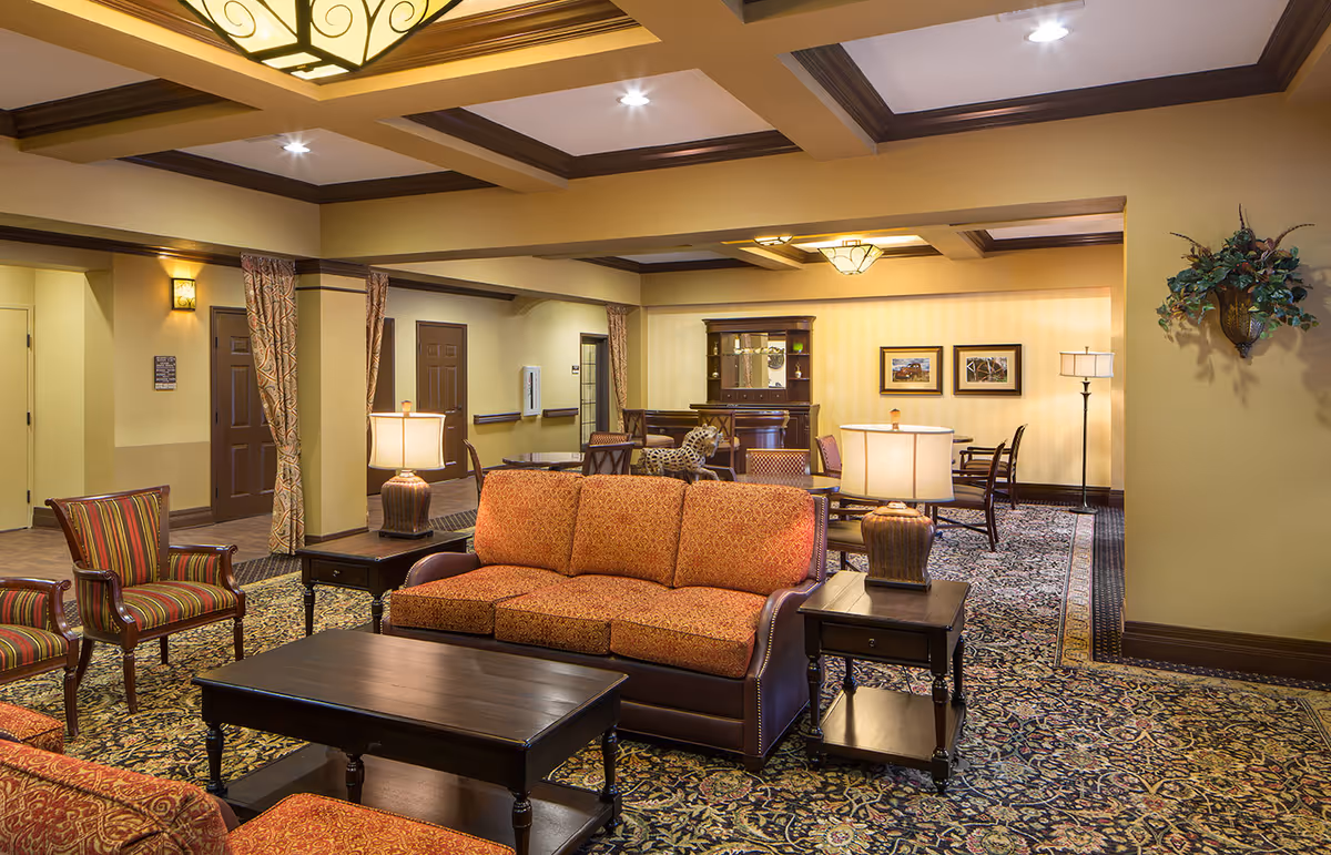 A spacious and warmly lit living room area in a senior living facility featuring patterned upholstered sofas and chairs, wooden coffee and side tables with lamps, a patterned carpet, and decorative wall art and plants. The room has a coffered ceiling with recessed lighting and a large light fixture.