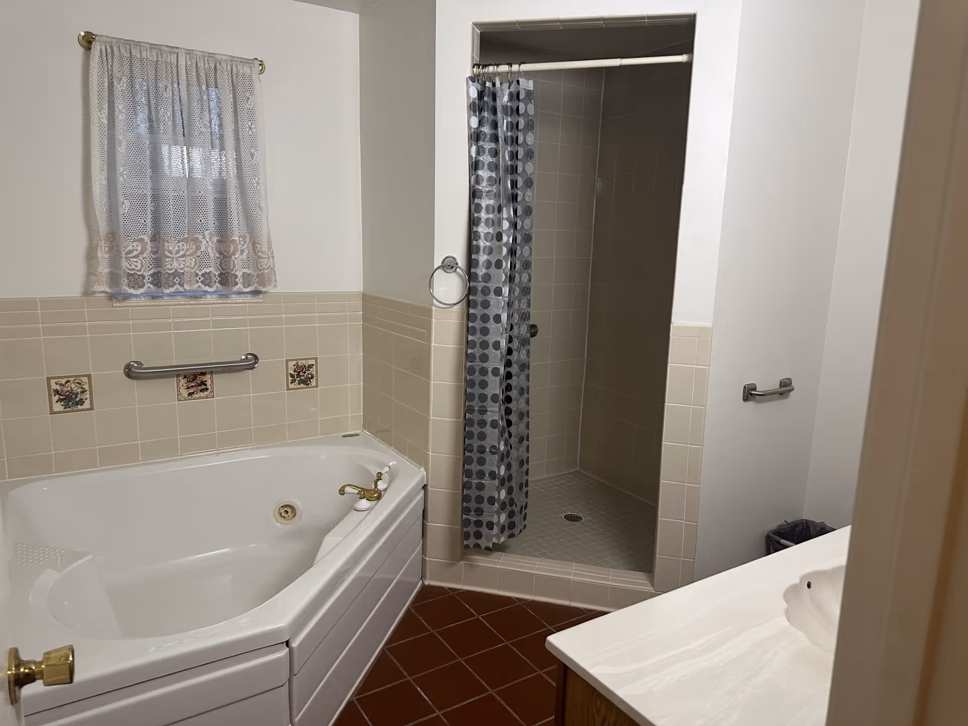 Bathroom with a corner bathtub featuring gold-colored fixtures, beige tiled walls with decorative floral tiles, a walk-in shower with a polka dot shower curtain, a small window with lace curtains, and a white sink countertop partially visible.