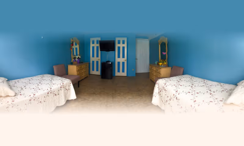 A bedroom with two single beds on opposite sides, each covered with white floral bedspreads. Between the beds are two wooden dressers with mirrors, each adorned with decorative items and a chair beside them. The walls are painted blue, and there is a TV mounted on the wall above a small black cabinet in the center. Two white doors with blue panels are visible in the background.