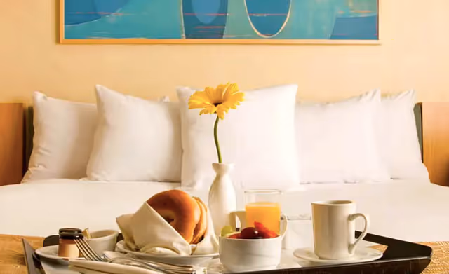 Breakfast tray with coffee, orange juice, a bagel and fruit set on a bed with white pillows and a single yellow flower in a vase.