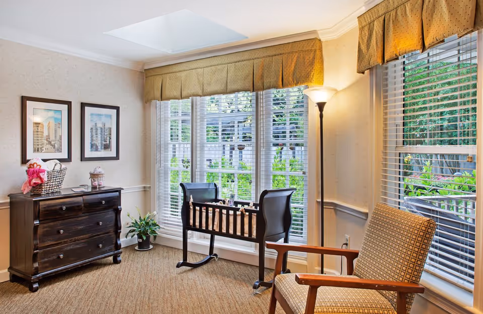 A cozy room with large windows covered by white blinds and green valances, featuring a dark wooden cradle, a patterned armchair, a tall floor lamp, a dark wooden dresser with a basket and decorative items on top, two framed pictures on the wall, and a small potted plant on the floor.