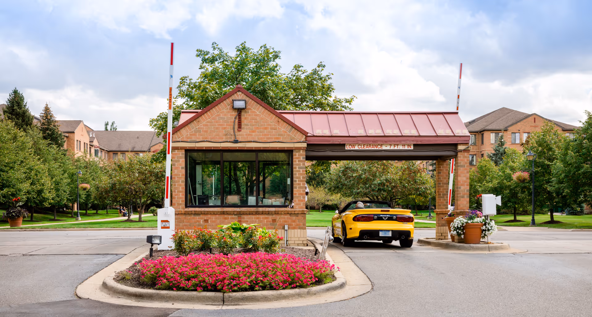 Entrance gatehouse of a senior living facility with a yellow convertible car passing through. The gatehouse is made of brick with a red metal roof and has a sign indicating low clearance of 7 feet 11 inches. There are landscaped flower beds with pink flowers and greenery around the entrance, and residential buildings and trees are visible in the background under a partly cloudy sky.
