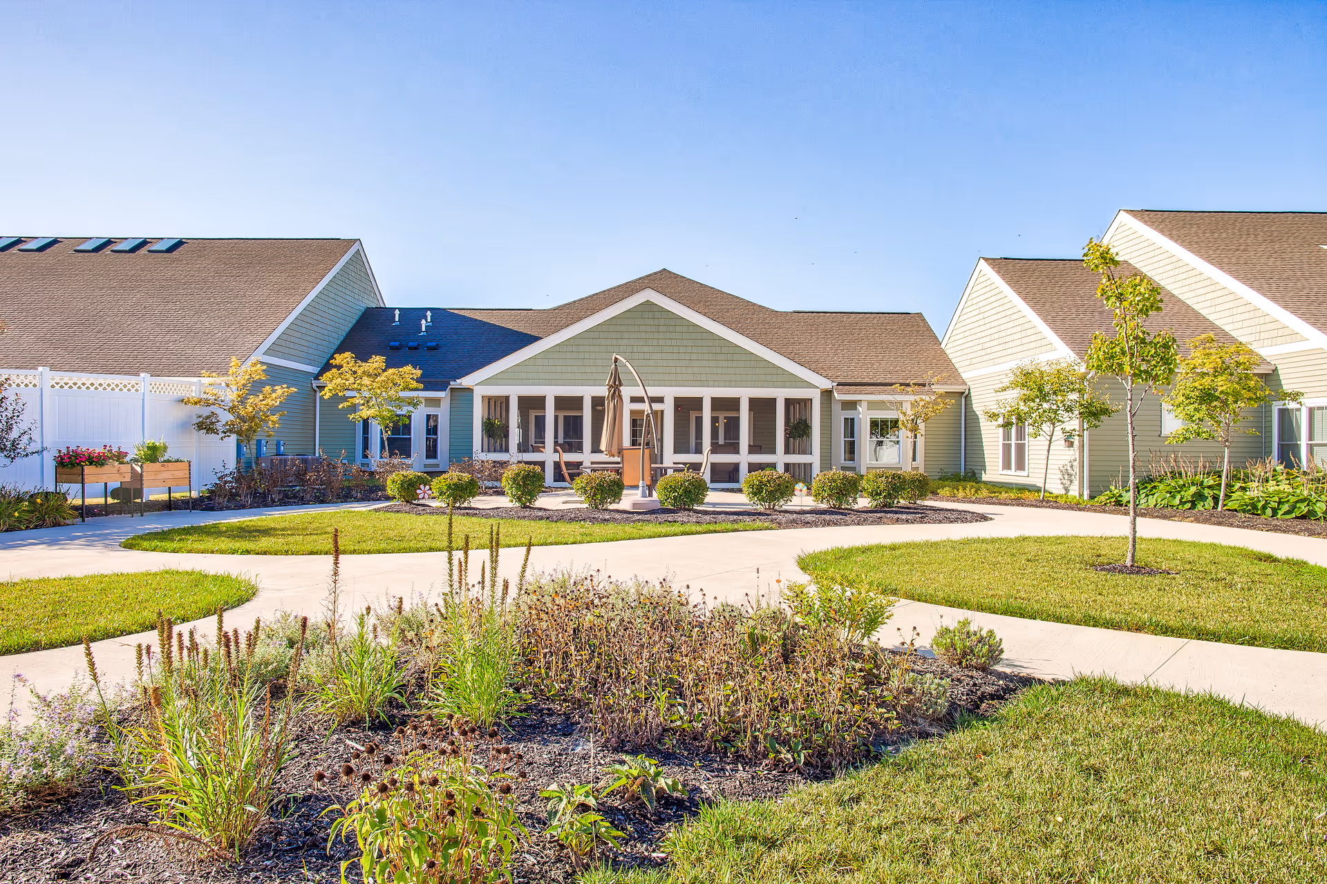 A bright outdoor courtyard area at Landings of Sidney featuring a paved walkway, landscaped garden beds with various plants and shrubs, small trees, and a building with a covered porch and seating area under a clear blue sky.