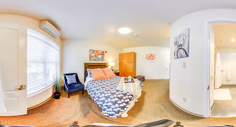 A bright and cozy bedroom with a bed covered in a patterned duvet and orange pillows. There is a blue armchair next to a large window with white blinds. A wooden wardrobe and a small ottoman are placed against the wall. The room has beige carpet and light-colored walls with colorful artwork. A doorway leads to a bathroom with a shower visible.
