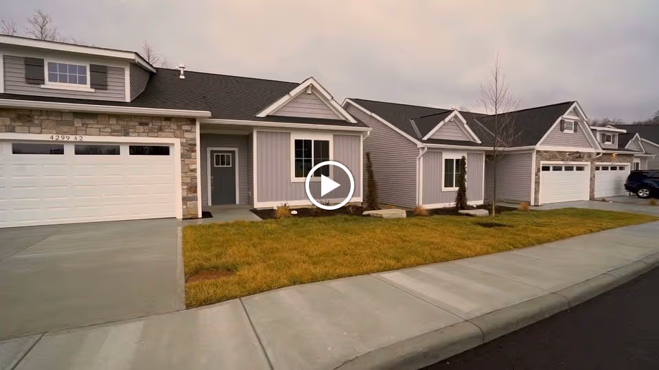Exterior view of a row of single-story residential units with attached garages, gray siding, stone accents, and small front lawns along a sidewalk under an overcast sky.