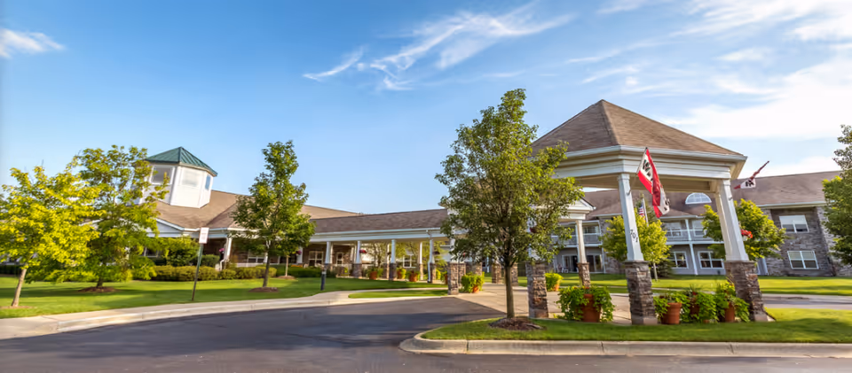 Exterior view of Independence Village of Oxford (Waterstone) showing a large building with a covered entrance supported by stone pillars, surrounded by green trees and a well-maintained lawn under a clear blue sky.