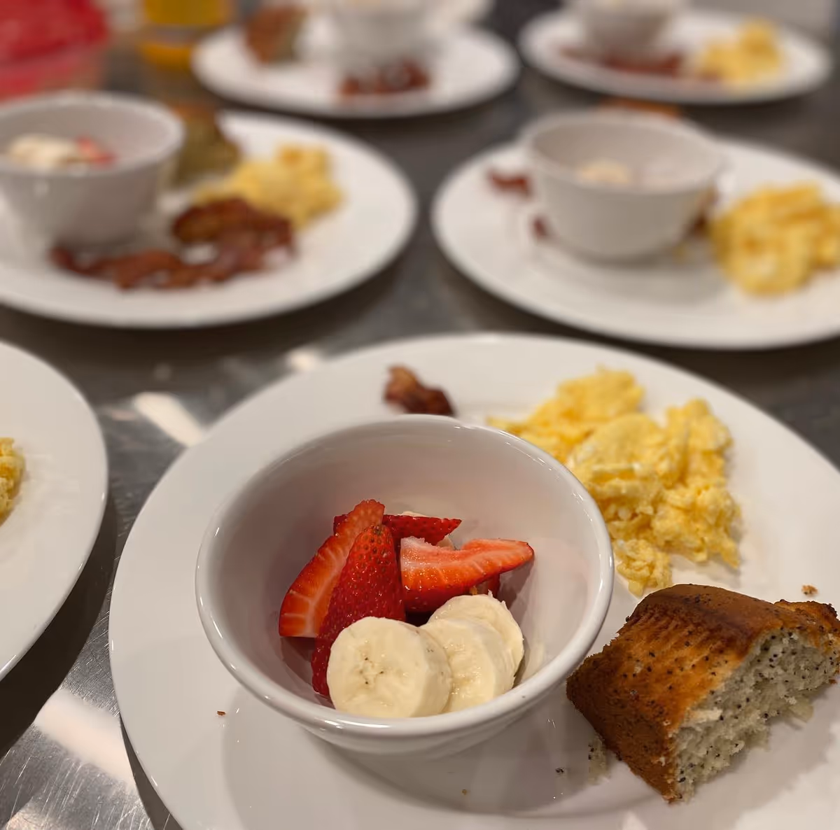 Plates of breakfast food on a table, each with scrambled eggs, a piece of poppy seed muffin, bacon, and a bowl containing sliced strawberries and bananas.