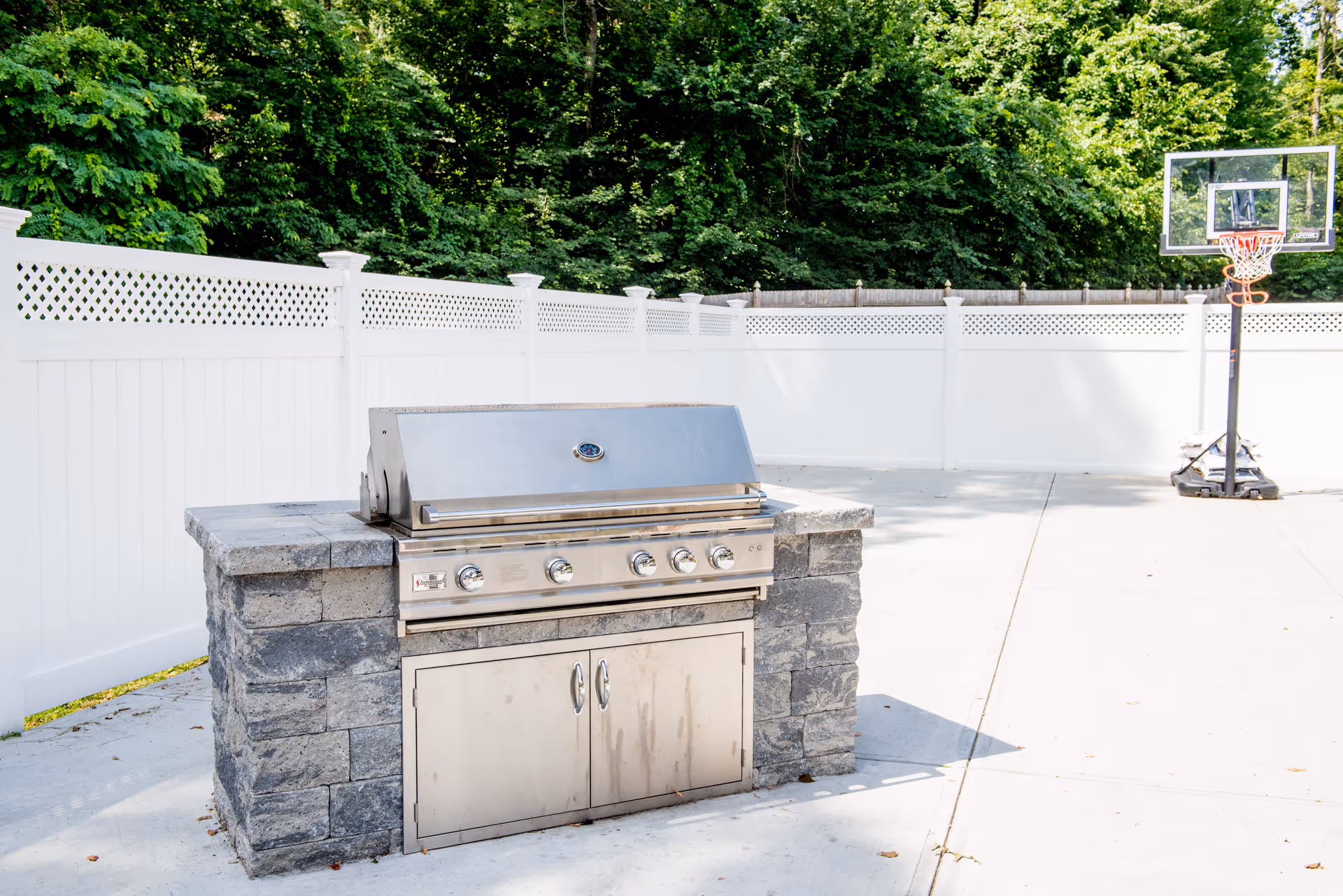 Outdoor area with a built-in stainless steel grill set in a stone countertop. The space is enclosed by a white fence with trees visible beyond it. A portable basketball hoop is positioned on the concrete ground nearby.