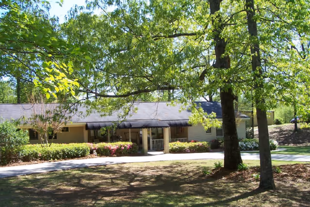 Single-story building of Bella Wood Senior Living surrounded by trees and greenery, with a pathway leading to the entrance under a black awning.