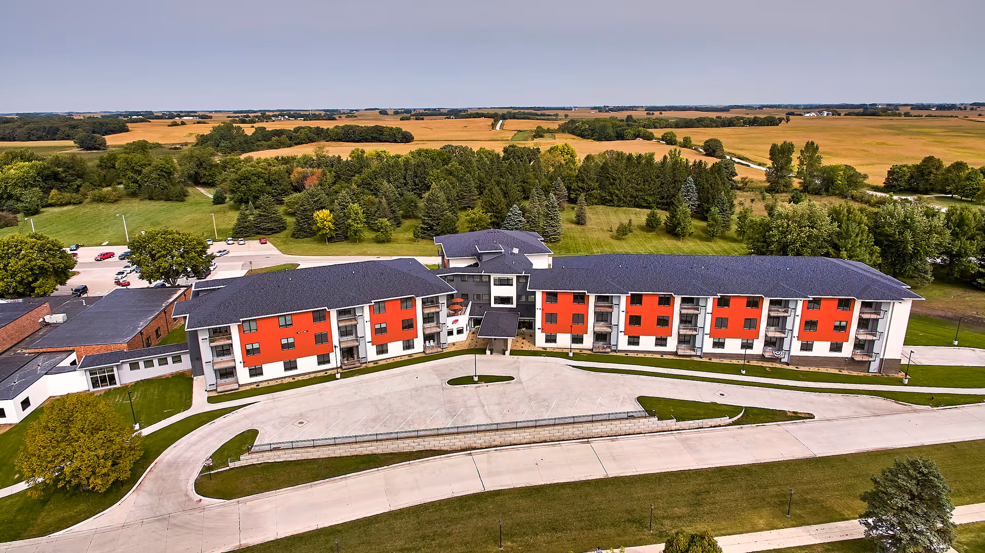 Aerial view of TimberCrest at Glen Oaks senior living facility showing a large, modern building with red and white exterior walls, surrounded by green lawns, trees, and parking areas, with expansive farmland in the background under a clear sky.