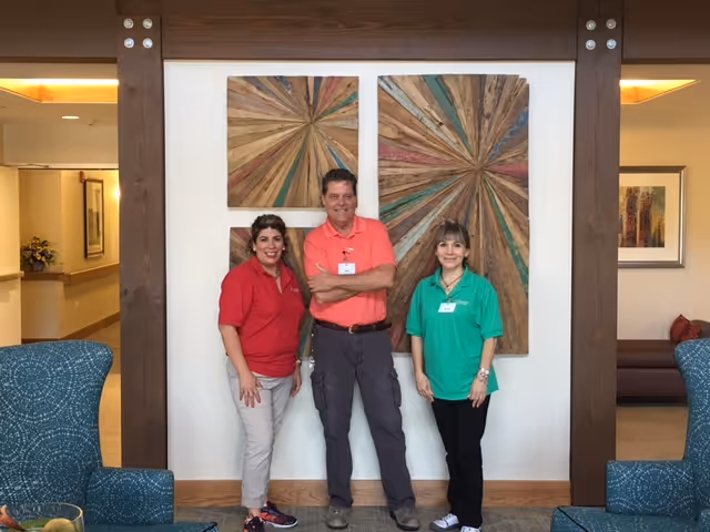 Three staff members standing and smiling inside a senior living facility lobby or common area, with wooden decorative wall art behind them and comfortable seating visible on the sides.