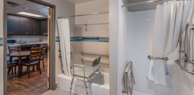 View of a bathroom with a shower and bathtub equipped with grab bars and a shower chair. The shower has a white curtain and a handheld showerhead. Through the open door, a dining area with a wooden table and chairs is visible.