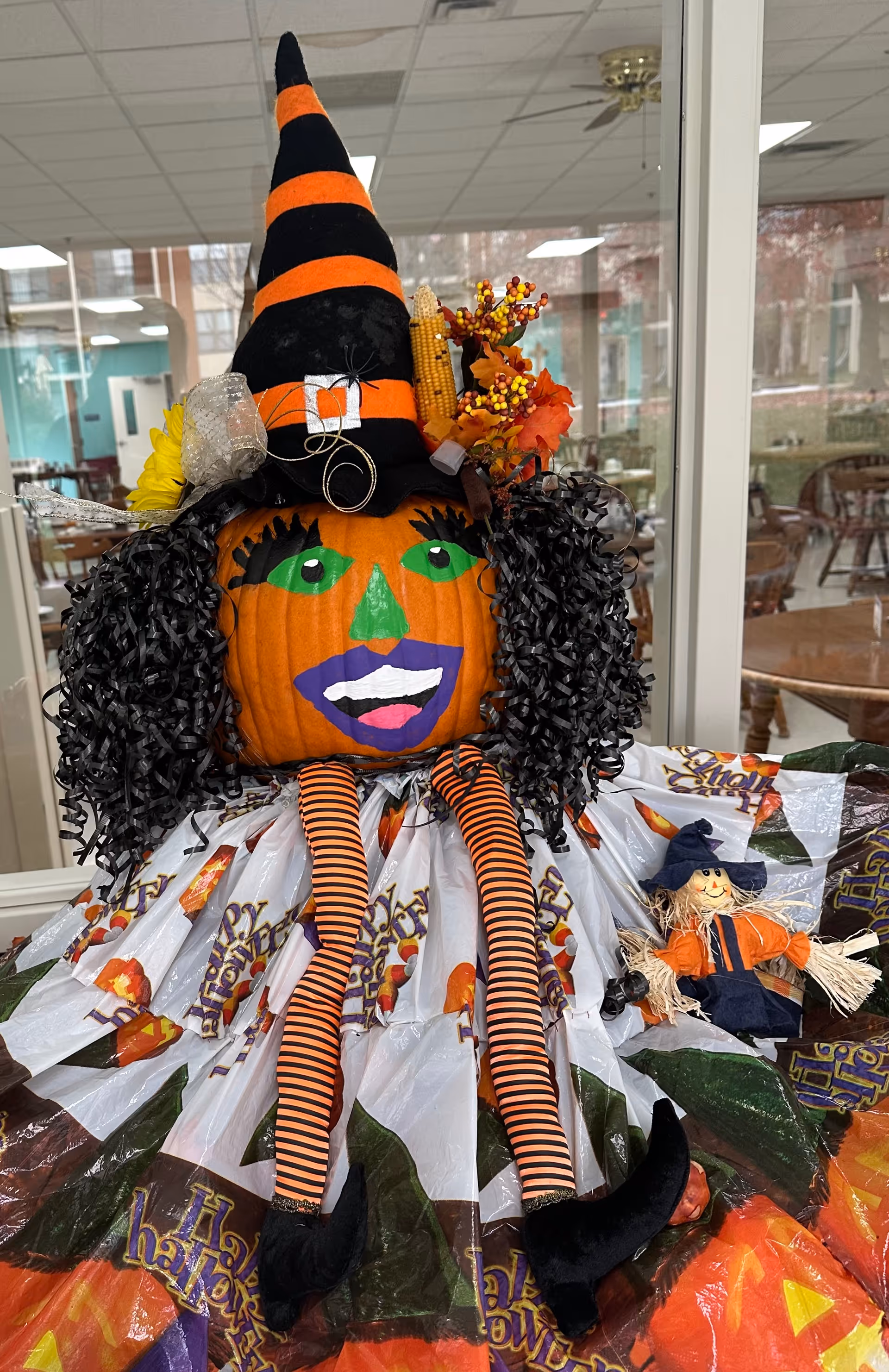 A Halloween-themed decoration featuring a pumpkin painted with a green nose, green eyes, purple lips, and black eyelashes. The pumpkin is wearing a black and orange striped witch hat adorned with autumn decorations like corn and leaves. It has curly black hair, striped orange and black legs, and black shoes. The pumpkin is seated on a table covered with a Halloween-themed plastic tablecloth. A small scarecrow doll dressed in orange and blue is placed next to the pumpkin. The background shows a room with tables and chairs and large windows.