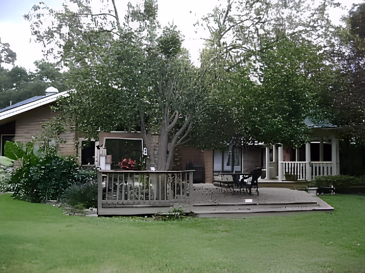 Wooden single-story house with a shaded deck and patio seating under large trees facing a green lawn.