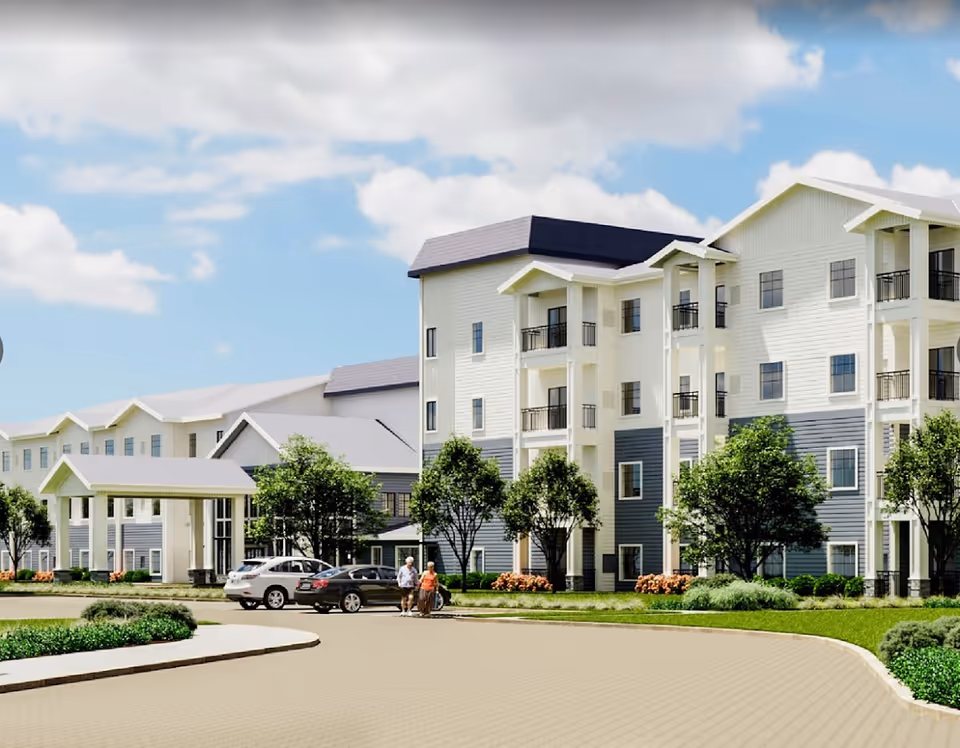 Exterior view of a multi-story senior living facility building with white and gray siding, balconies, and a covered entrance. There are trees, shrubs, and flowers around the building, a paved driveway with parked cars, and two elderly people walking near the entrance under a partly cloudy sky.