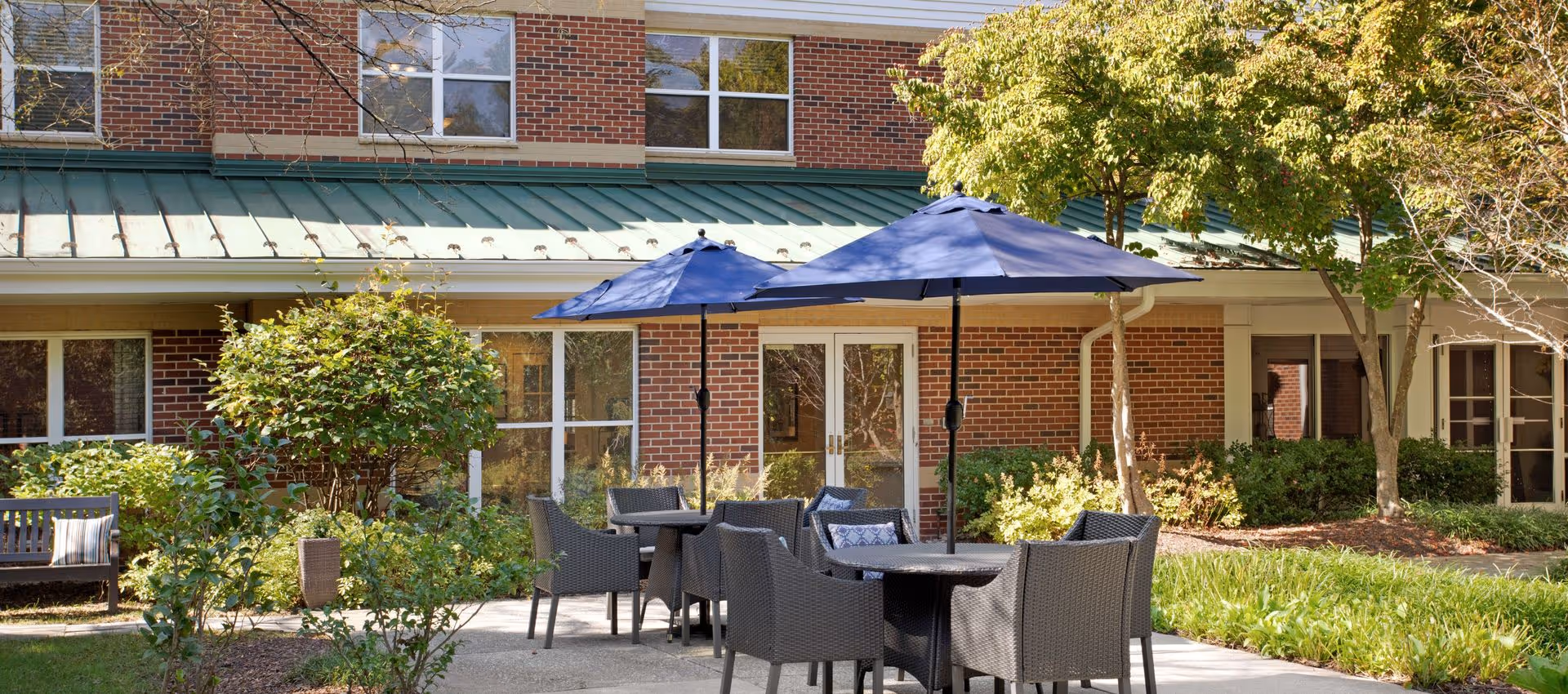 Outdoor patio area at Brighton Gardens of Columbia with black wicker tables and chairs under blue umbrellas, surrounded by green bushes and trees, in front of a brick building with windows and glass doors.