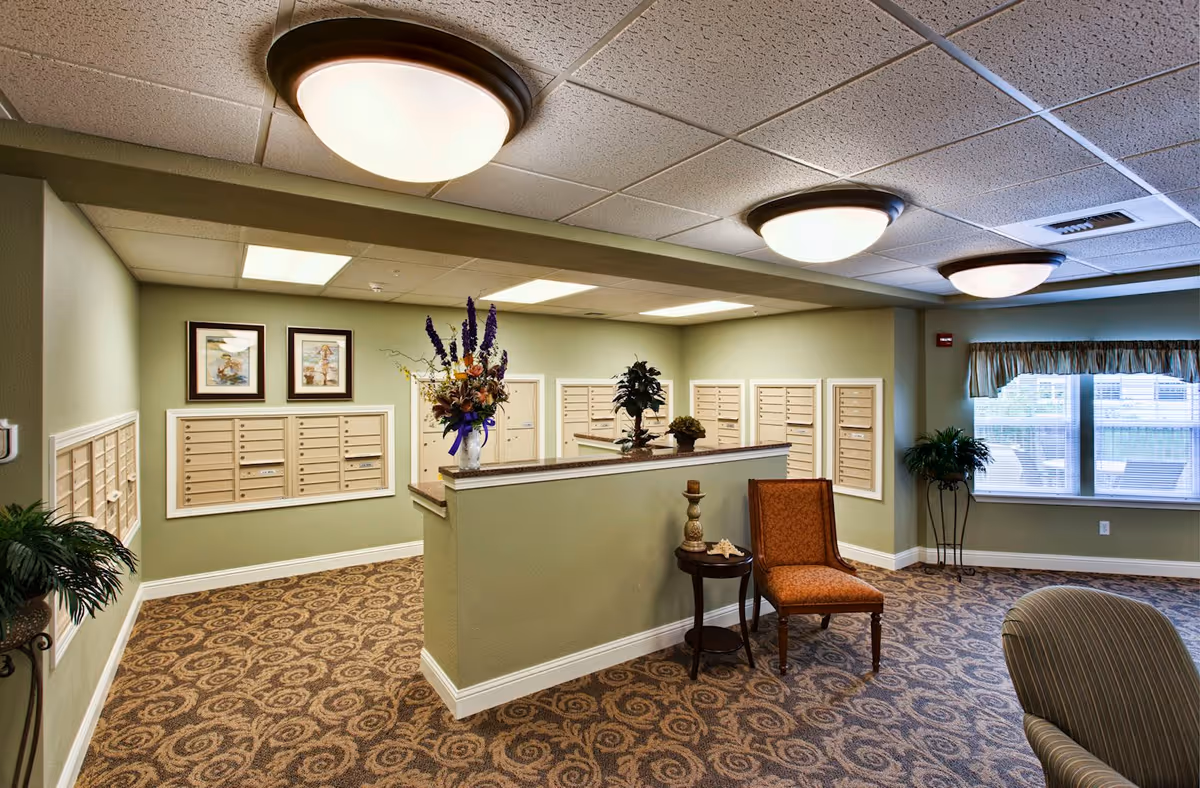 Interior view of a senior living community mailroom with multiple mailboxes mounted on light green walls, a small table with decorative items, an orange upholstered chair, patterned carpet, ceiling lights, framed pictures, and a window with a valance.