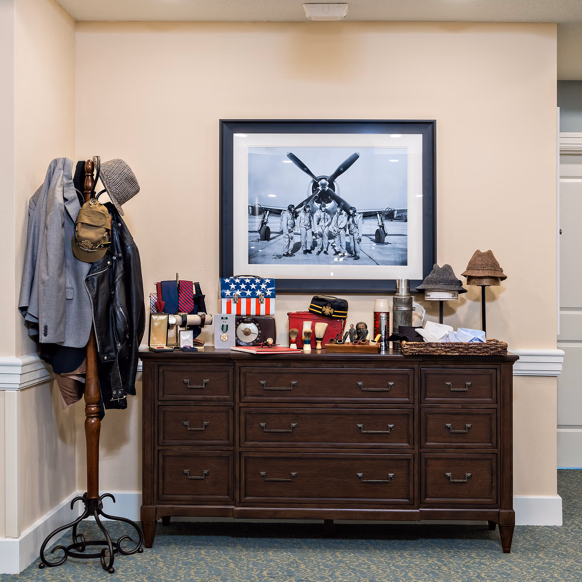 A wooden dresser with multiple drawers is placed against a beige wall. On top of the dresser are various vintage items including shaving brushes, a radio, hats, and a small American flag-themed box. Above the dresser hangs a black and white framed photograph of six men in military uniforms standing in front of an airplane propeller. To the left of the dresser is a wooden coat rack holding a gray jacket, a black leather jacket, and two hats. The floor is carpeted in a blue patterned design.