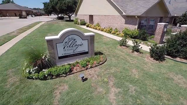 Lawn and entrance sign for 'Villagio of Plano' in front of a brick building with a walkway and landscaping.