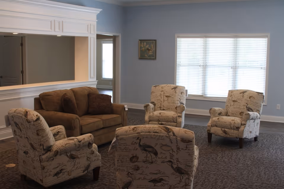 A cozy living room area with a brown sofa and four beige armchairs featuring bird and script patterns. The room has light blue walls, a large window with white blinds, and a framed picture on the wall.