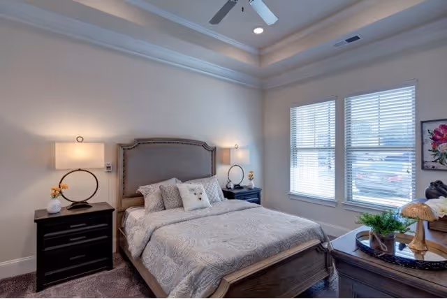 A well-lit bedroom with a large bed featuring a padded headboard, two bedside tables each with a lamp, a ceiling fan, and two large windows with blinds letting in natural light. The room has neutral-colored walls and carpeted flooring.
