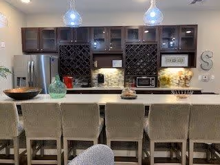 Modern kitchen with a large island countertop featuring six upholstered bar stools. The kitchen has dark wood cabinets, stainless steel refrigerator, built-in wine racks, and pendant lights hanging above the island. Various decorative items and kitchen appliances are placed on the countertop and shelves.