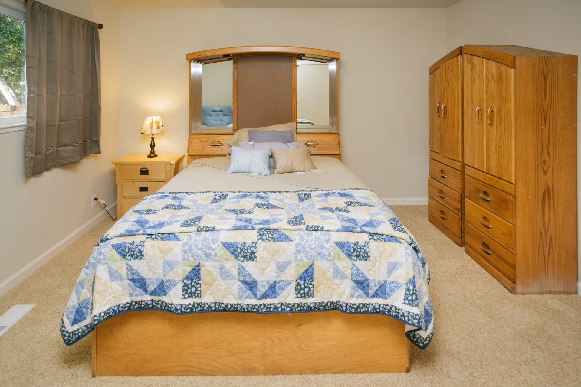 A neatly made bed with a wooden headboard featuring three mirrors, covered with a quilt in blue, beige, and white patterns. There are several pillows on the bed. To the left of the bed is a wooden nightstand with a lamp on it, and to the right are two wooden dressers. A window with gray curtains is on the left wall, and the room has beige carpet flooring and light-colored walls.