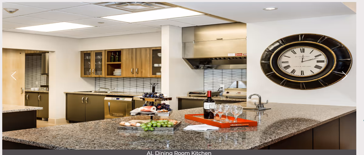 Modern kitchen area with granite countertops, wooden cabinets, a wall clock, and a tray with wine bottle and glasses on the counter. The kitchen features stainless steel appliances and a tiled backsplash.