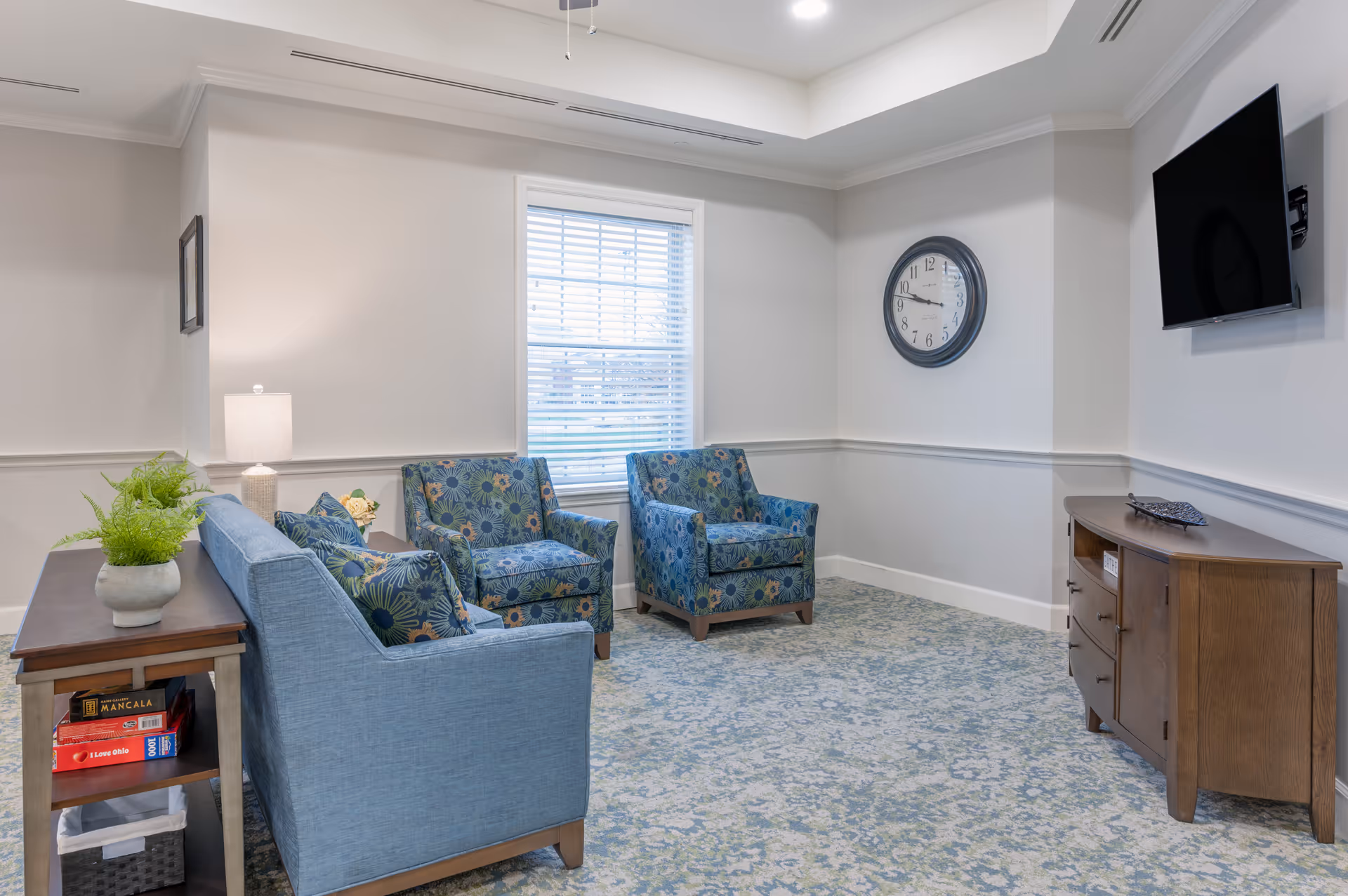 A cozy living room area with a blue sofa and two patterned armchairs arranged around a window with blinds. There is a wooden side table with a lamp and a plant, and a wooden cabinet with a flat-screen TV mounted above it. A round wall clock is mounted on the wall, and the room has a patterned carpet and neutral-colored walls.