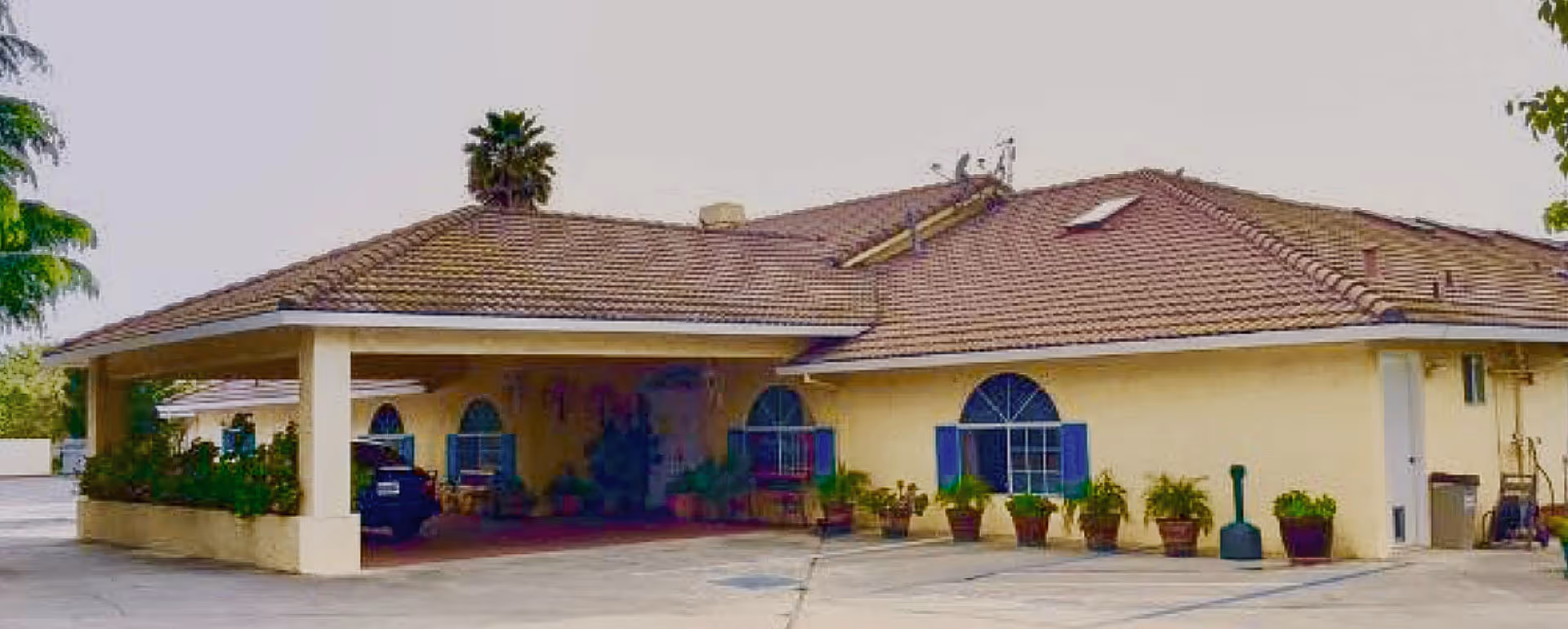 Exterior view of Whispering Pines Inn, a single-story building with a tiled roof, arched windows with blue shutters, and several potted plants along the front. There is a covered entrance area with a parked vehicle and surrounding greenery.