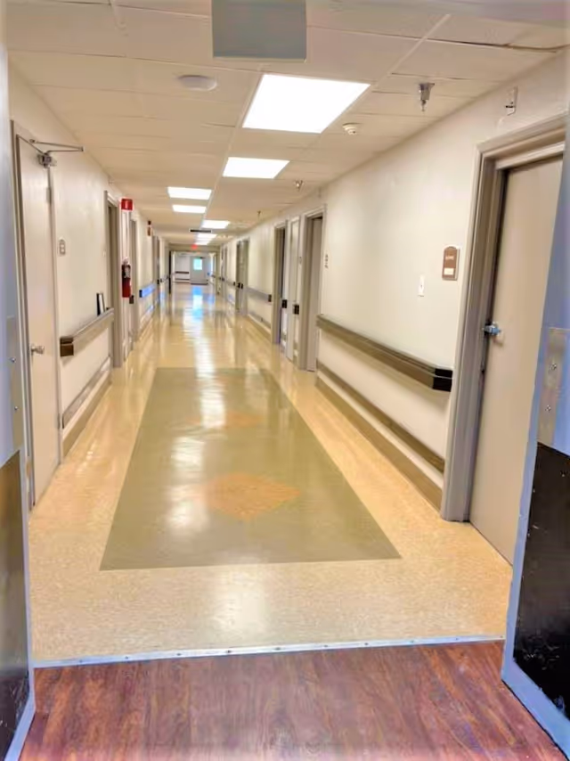 A long, clean hallway inside a senior care facility with beige and green patterned flooring, white walls, multiple closed doors on both sides, handrails along the walls, and ceiling lights.