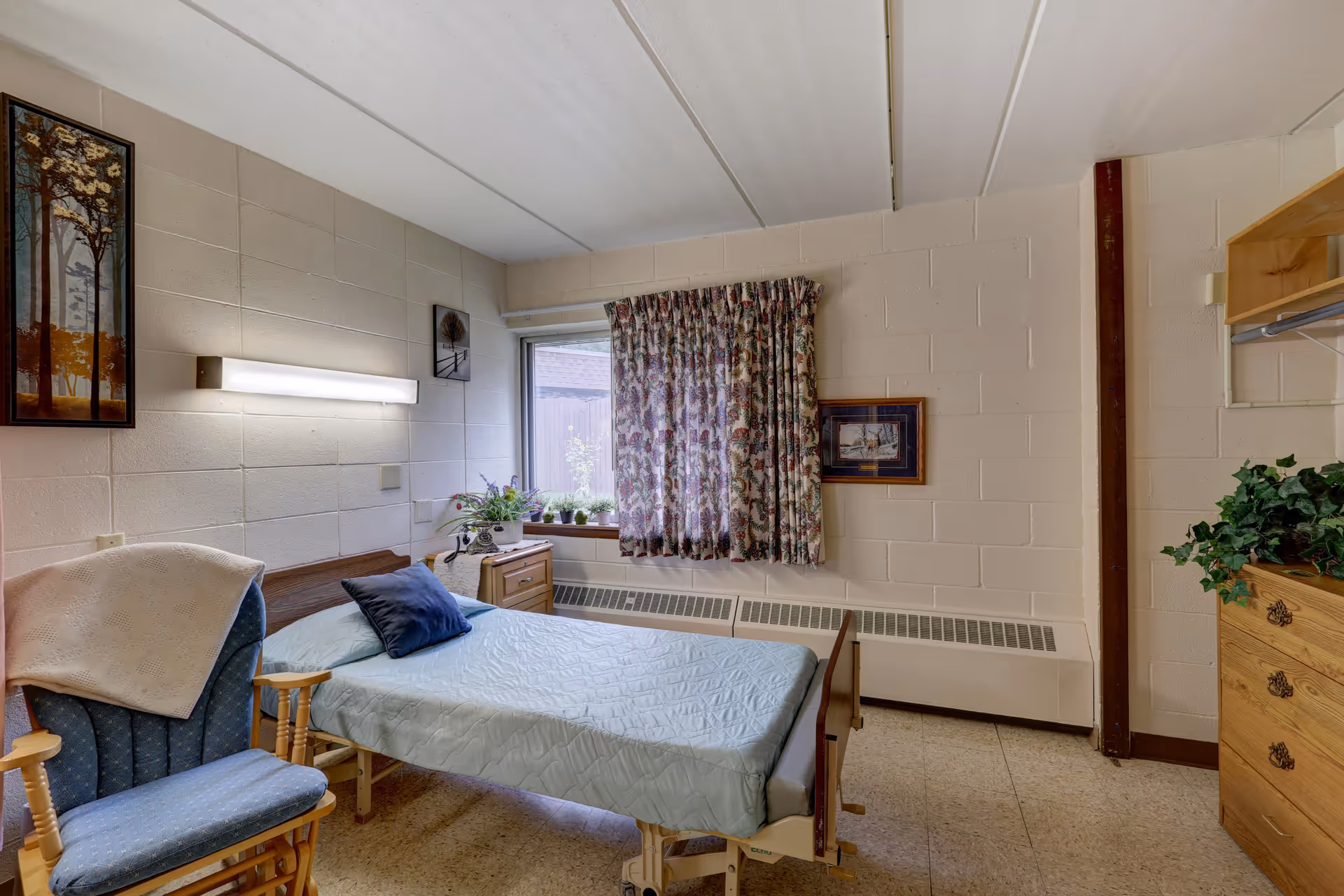 A simple bedroom in a senior living facility with a single bed covered in a light blue quilt, a blue cushioned rocking chair with a white blanket draped over it, a wooden nightstand with potted plants, a window with floral curtains, and framed artwork on the walls.